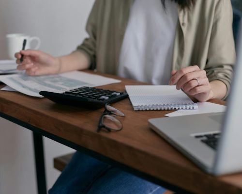 pexels-photo-6963857-6963857 A woman manages finances at home, using a laptop and calculator on a wooden desk.
